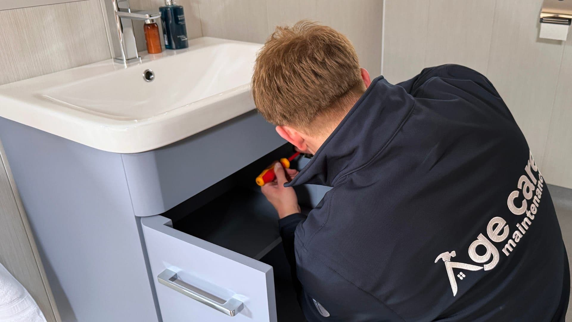 An Age Care Maintenance handyman working under a bathroom sink