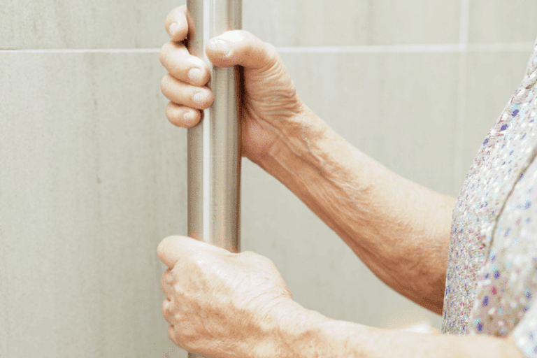 An older woman’s hands holding a grab rail in a tiled bathroom