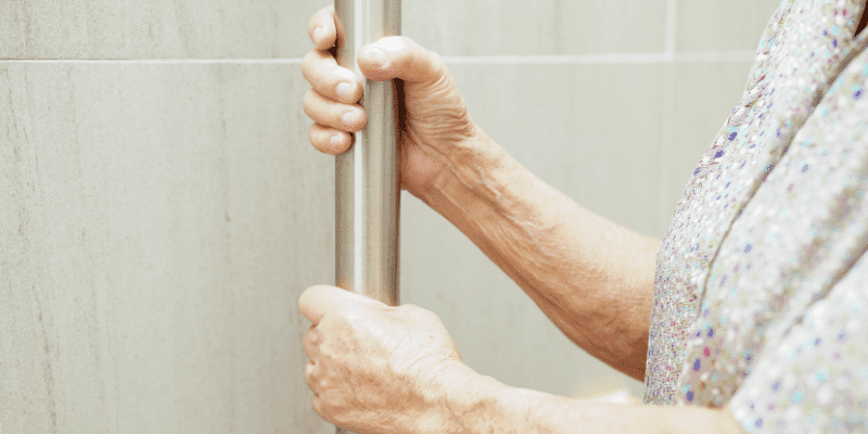 An older woman’s hands holding a grab rail in a tiled bathroom